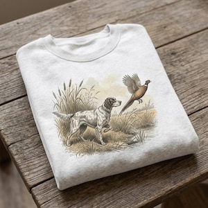May include: A light gray crewneck sweatshirt featuring a detailed illustration of a hunting dog standing in tall grass and reeds, watching a pheasant in flight. The graphic is rendered in muted earth tones, depicting a natural outdoor scene.