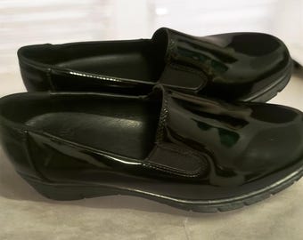 May include: A pair of black slip-on shoes with a glossy patent finish. These shoes feature a rounded toe, elastic side gussets for a flexible fit, and a thick, textured black rubber sole that provides a stable base for the footwear.