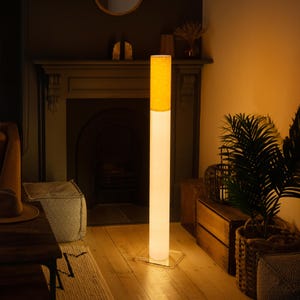 May include: A modern floor lamp with a cylindrical design. The lamp features a bright yellow lampshade atop a long, white column. The base is a clear, square shape. The lamp is in a room with a fireplace and wooden furniture.