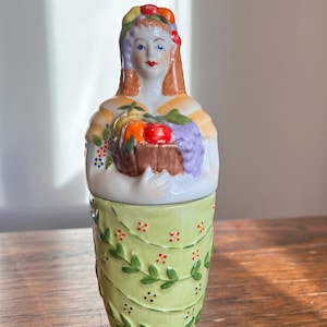 May include: A ceramic figurine featuring a person with a fruit-adorned headpiece holding a basket of colourful fruit. The figure wears a light yellow top and a light green skirt decorated with a repeating vine pattern and small red and blue floral accents.