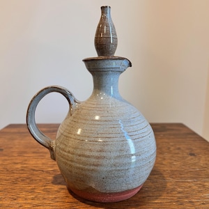 May include: A handcrafted ceramic jug with a handle and a spout. The jug is mainly beige with blue details and a red base. A separate, brown stopper sits on top. The jug is likely used for serving liquids.