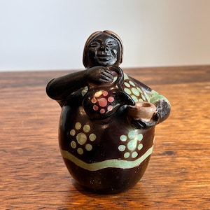 May include: A dark brown ceramic figurine of a woman holding a jug and a small bowl. The figurine has a rounded body and is decorated with green and pink polka dots. The woman's face has a smiling expression.