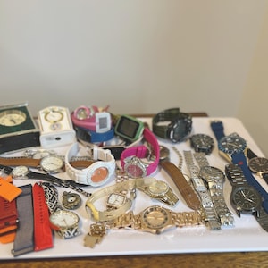 May include: A collection of various wristwatches and two small desk clocks. The assortment includes analogue and digital timepieces with straps in colours such as orange, red, white, gold, silver, black, and blue, featuring diverse face designs.
