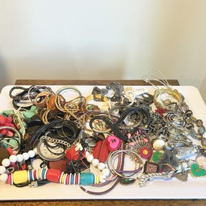 May include: A large collection of assorted fashion jewellery including beaded bracelets, metal bangles, charm necklaces, and layered chains in various colours like red, yellow, blue, and silver, all piled together on a white rectangular tray.