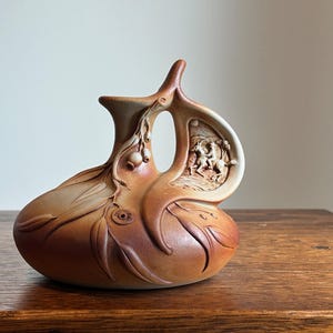 May include: A tan ceramic vase with an elaborate design. The vase has a rounded base, a handle with a carved relief of figures, and a spout. The surface is decorated with leaf and floral patterns, and the style is artistic.