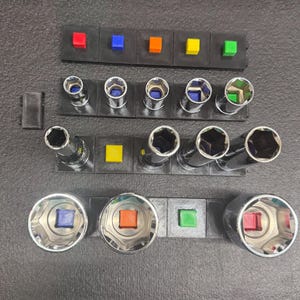 May include: A set of metal socket tools organized on black plastic holders. The arrangement includes various cylindrical sockets of different sizes, each featuring a small, colored square insert in red, blue, orange, yellow, or green.