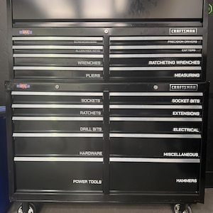 May include: A black Craftsman rolling tool chest with multiple drawers, each labeled with white text including Screwdrivers, Allen/Hex Keys, Wrenches, Pliers, Precision Drivers, Cutters, Ratcheting Wrenches, Measuring, Sockets, Socket Bits, Ratchets, Extensions, Drill Bits, Electrical, Hardware, Miscellaneous, Power Tools, and Hammers.