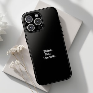 May include: Black smartphone case with the text "Think. Plan. Execute." in white. The phone case has a glossy finish and a camera module with four lenses. The phone is resting on a white surface, with white flowers in the background.