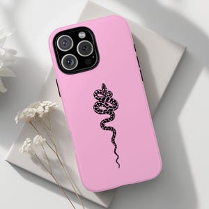 Könnte beinhalten: Eine rosa Handyhülle mit einem schwarzen Schlangenmuster. Die Hülle wird auf einer weißen Oberfläche mit weißen Blumen im Hintergrund gezeigt. Die Handyhülle hat ein schwarzes Kameramodul.