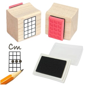 May include: Two wooden stamp blocks with red rubber inserts. One block has a grid pattern and the other has a "Cm" chord diagram for ukulele. A separate ink pad is also included.