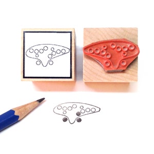 May include: Two wooden blocks with a black outline of a tin whistle on one block and a red rubber stamp of a tin whistle on the other block. A pencil is lying on a white surface next to a black outline of a tin whistle.