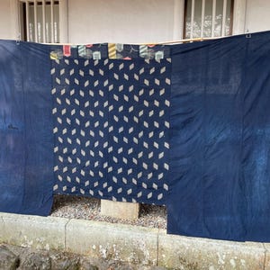May include: A large indigo-dyed textile, featuring a central panel with white diamond patterns on a dark blue backdrop. The top section has a patchwork design with colourful geometric shapes. The fabric is displayed outdoors, possibly a traditional craft or decorative item.