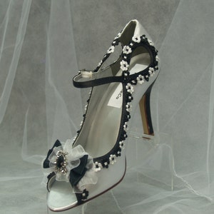 May include: A silver satin high heel shoe with a black bow and floral embellishments. The shoe has a peep toe and a high heel.