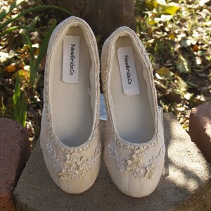 Puede incluir: Un par de zapatos de novia de sat&eacute;n color marfil con delicado bordado floral y una etiqueta NewBrideCo. Los zapatos est&aacute;n sobre una superficie de piedra.