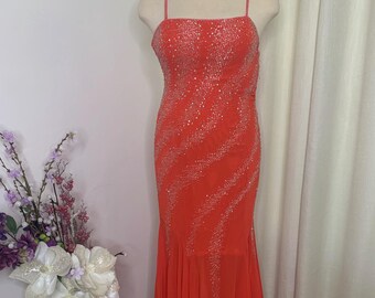 Könnte beinhalten: Ein leuchtend orangefarbenes Abendkleid mit einem figurbetonten Oberteil und einem fließenden, mehrlagigen Rock. Das Kleid hat dünne Spaghettiträger und ist mit glitzernden silbernen Akzenten verziert. Das Kleid wird auf einer Schaufensterpuppe präsentiert.