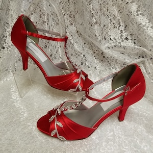 May include: A pair of red satin bridal shoes with a t-strap and rhinestone embellishments. The shoes have a delicate leaf design and a peep toe.