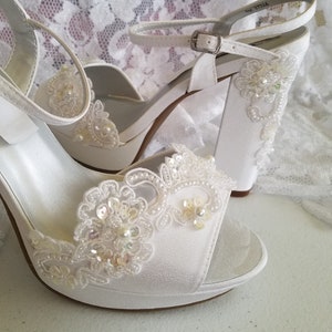 May include: White satin bridal shoes with a chunky heel and delicate lace embellishments. The shoes have a peep toe and an ankle strap.