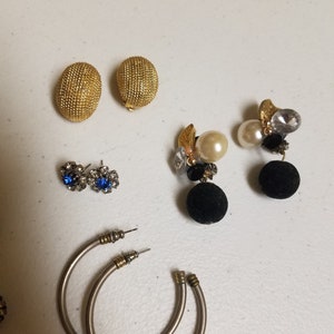 May include: Assortment of earrings, including a pair of gold hoop earrings, a pair of gold ball earrings, a pair of blue rhinestone earrings, and a pair of black and gold dangle earrings with pearls.