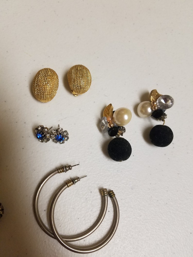 May include: Assortment of earrings, including a pair of gold hoop earrings, a pair of gold ball earrings, a pair of blue rhinestone earrings, and a pair of black and gold dangle earrings with pearls.