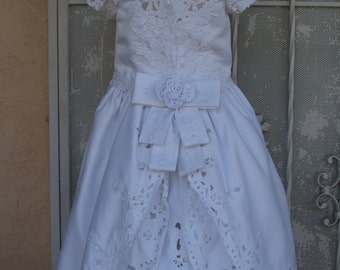Puede incluir: Vestido blanco de encaje con falda de satén y un gran lazo en la cintura. El vestido tiene un borde de encaje festoneado y un delicado diseño floral.