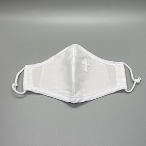 May include: White face mask with a small embroidered cross on the front. The mask has adjustable white ear loops and a white trim. The mask is made of a soft, white fabric.