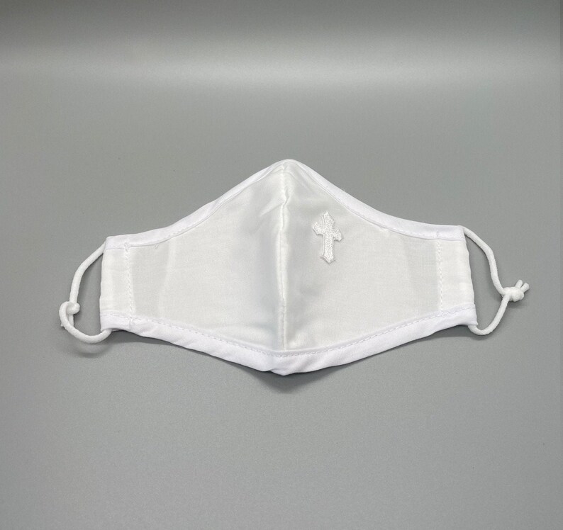 May include: White face mask with a small embroidered cross on the front. The mask has adjustable white ear loops and a white trim. The mask is made of a soft, white fabric.