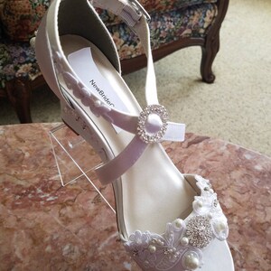 Pode incluir: Sapatos de noiva de cetim branco com um delicado adorno de renda e p&eacute;rolas. Os sapatos t&ecirc;m um salto de cunha e uma tira de tornozelo com uma fivela de strass. O r&oacute;tulo no sapato diz "NewBrideC".