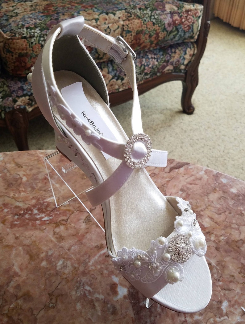 Pode incluir: Sapatos de noiva de cetim branco com um delicado adorno de renda e p&eacute;rolas. Os sapatos t&ecirc;m um salto de cunha e uma tira de tornozelo com uma fivela de strass. O r&oacute;tulo no sapato diz "NewBrideC".