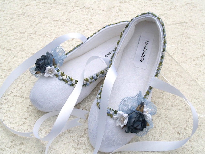 May include: White satin bridal shoes with blue and green floral embroidery and lace accents. The shoes have satin ribbons and a label that reads "NewBrideCo."