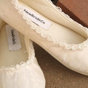 May include: A pair of white satin bridal slippers with delicate lace trim. The slippers have a label that reads "NewBrideCo".