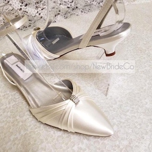 May include: A pair of white satin bridal shoes with a low heel and a rhinestone embellishment. The shoes have ankle straps and are on a white lace background.