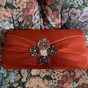 May include: A rectangular rust-colored satin clutch bag featuring a central decorative cluster of three fabric flowers. The flowers are embellished with metallic beads, sequins, and clear crystals, with two dark brown fabric leaves at the base.