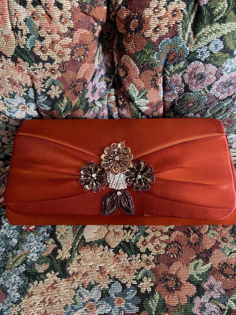 May include: A rectangular rust-colored satin clutch bag featuring a central decorative cluster of three fabric flowers. The flowers are embellished with metallic beads, sequins, and clear crystals, with two dark brown fabric leaves at the base.