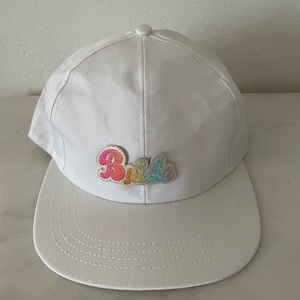Könnte beinhalten: Weiße Baseballkappe mit einem regenbogenfarbenen gestickten "Bride"-Aufnäher.