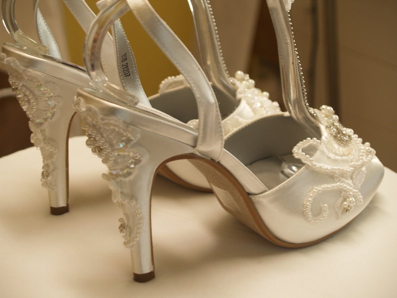 May include: A pair of white satin high-heeled shoes featuring an ankle strap and a T-strap design. The shoes are decorated with intricate white beadwork, sequins, and embroidery along the heel, sides, and front, creating a textured pattern.