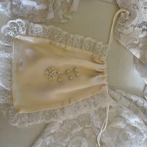 May include: A small, off-white satin drawstring bag with a lace trim and pearl embellishments. The bag is tied with a white cord.