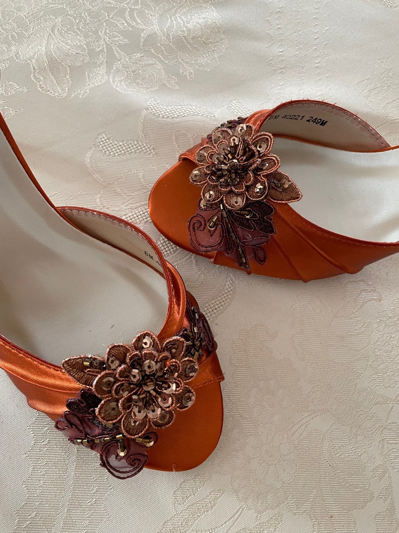 May include: A pair of burnt orange satin open-toe shoes featuring a decorative floral appliqu&eacute; on the vamp. The floral embellishment is crafted from dark brown fabric petals, metallic sequins, and beads. The interior lining is white.