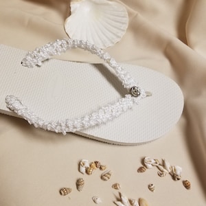 May include: White flip flops with a white crocheted strap and a small silver embellishment. The flip flops are on a beige fabric background with small seashells scattered around them.