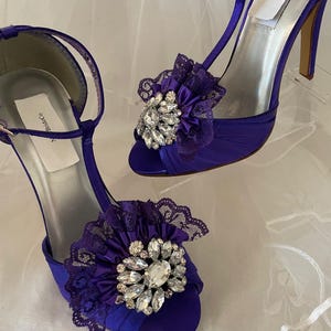 May include: A pair of elegant, purple satin high-heeled shoes. Each shoe features a decorative floral embellishment with lace trim and sparkling crystal accents. The shoes have a T-strap design and a slender heel.