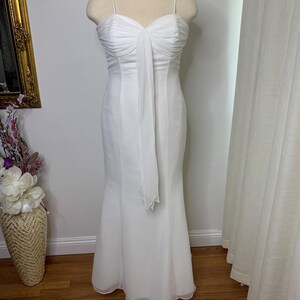 May include: A white formal gown with a sweetheart neckline and spaghetti straps. The bodice features a ruched design, and a flowing fabric panel drapes down the front. The dress has a fitted silhouette that flares out at the hemline. The gown is displayed on a mannequin.