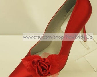 Puede incluir: Zapatos de tacón alto de satén rojo con un adorno de flor de rosa roja. Los zapatos tienen una punta puntiaguda y un tacón fino.