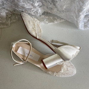May include: Ivory satin bridal flats with delicate lace and pearl embellishments. The shoes feature an ankle strap and a low heel. The label on the insole reads "New Bride Co.". A white lace fabric is draped in the background.
