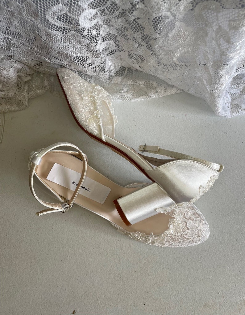 May include: Ivory satin bridal flats with delicate lace and pearl embellishments. The shoes feature an ankle strap and a low heel. The label on the insole reads "New Bride Co.". A white lace fabric is draped in the background.