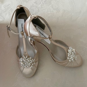 May include: A pair of ivory satin bridal shoes with a t-strap design and pearl embellishments. The shoes have a low heel and are labeled "NewBride".