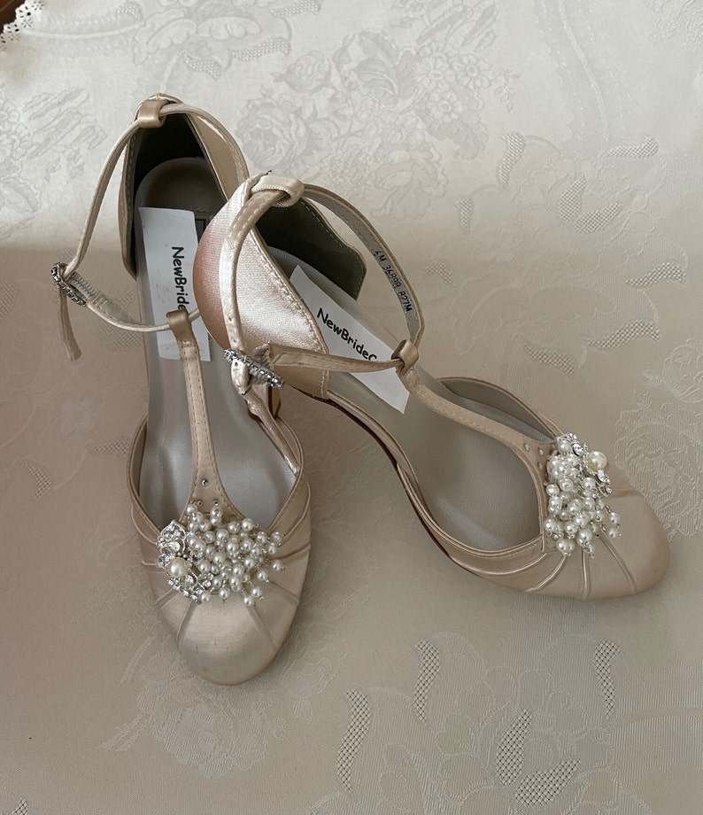 May include: A pair of ivory satin bridal shoes with a t-strap design and pearl embellishments. The shoes have a low heel and are labeled "NewBride".
