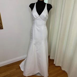 May include: A white halter wedding dress on a black mannequin. The dress has a V-neckline with a beaded embellishment at the waist. The skirt flares out slightly at the bottom, and the dress has a small train.