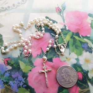May include: A white pearl rosary with a silver crucifix and a silver chain. The rosary is laid out on a floral patterned surface with a US quarter for scale.