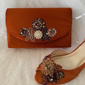May include: A burnt orange satin clutch bag and a matching peep-toe shoe. The clutch features a central floral appliqu&eacute; with sequins, beads, and crystals. The shoe has a similar floral embellishment on the toe, featuring brown and gold tones.