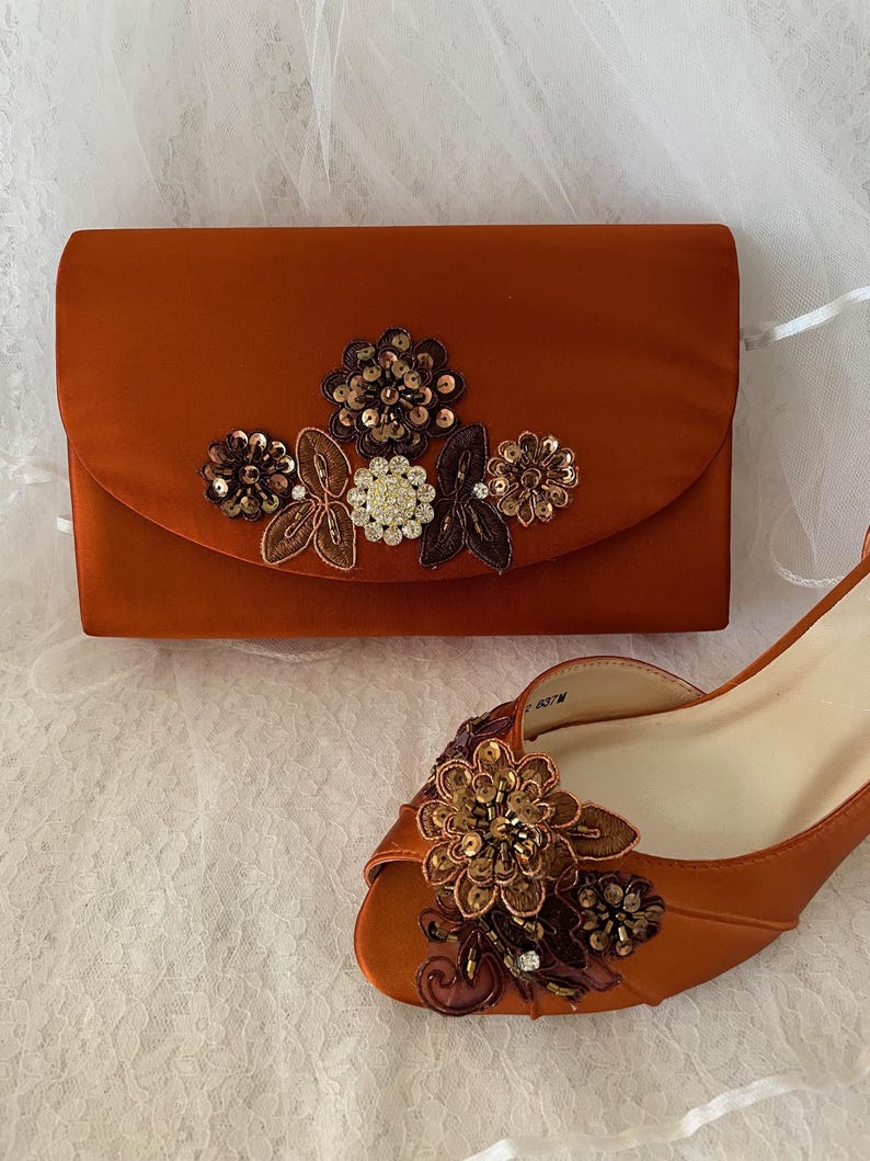 May include: A burnt orange satin clutch bag and a matching peep-toe shoe. The clutch features a central floral appliqu&eacute; with sequins, beads, and crystals. The shoe has a similar floral embellishment on the toe, featuring brown and gold tones.