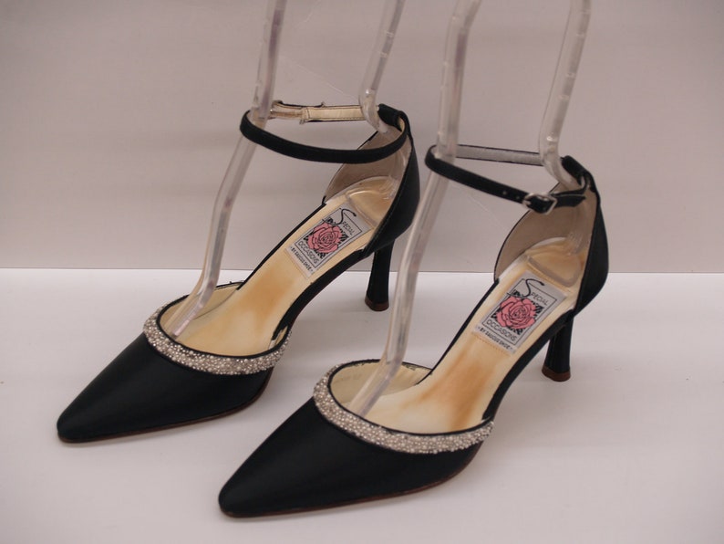 May include: A pair of black satin heeled shoes with ankle straps and a rhinestone trim. The shoes have a pointed toe and a delicate heel.